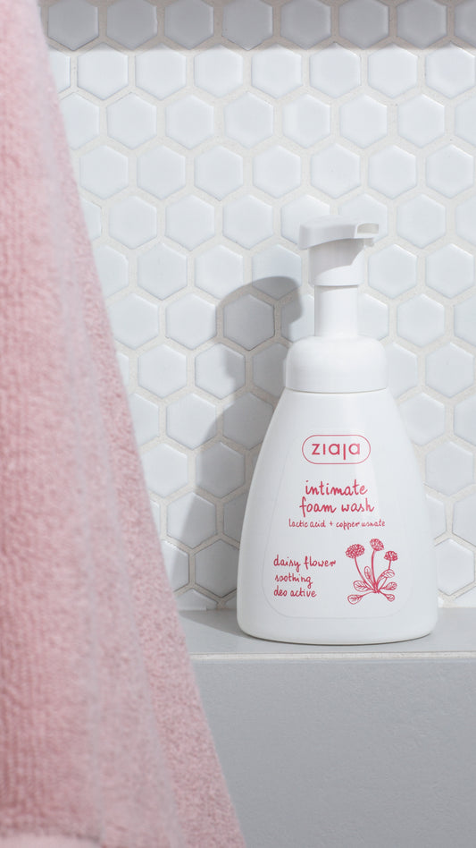 Ziaja intimate foam wash bottle on a bathroom shelf with pink towel and tiled wall.