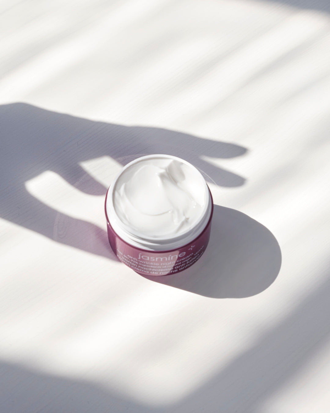 A small open jar of white cream or balm with a dark pink label sits on a white surface in bright sunlight with strong shadows cast across it.