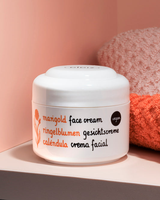 A white jar of marigold face cream by Ziaja styled on a pink surface with a textured pink towel in the background.