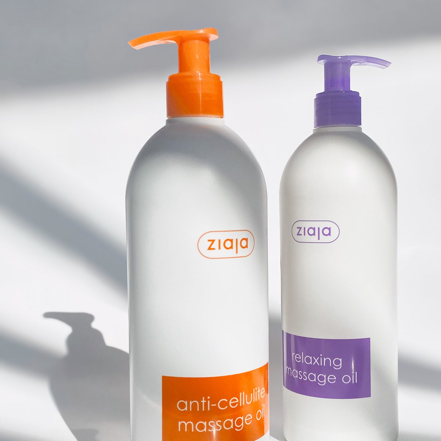 Two bottles of Ziaja massage oil, one with an orange pump and the other with a purple pump, on a neutral background.