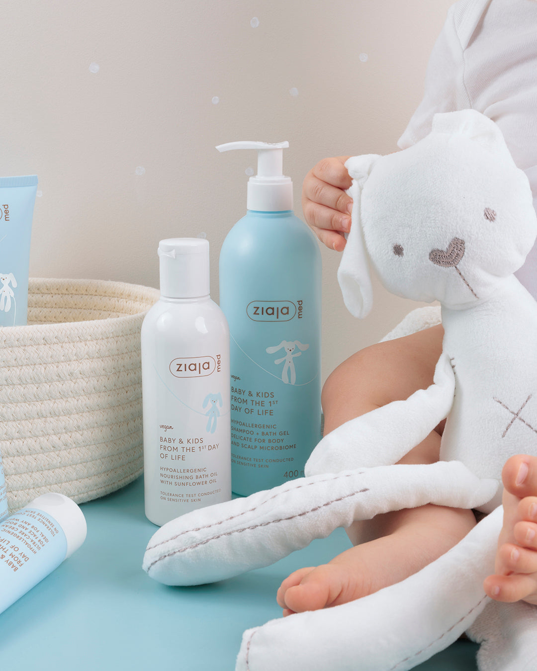 Ziaja Med Baby & Kids Nourishing Bath Oil (with sunflower oil) bottle shown next to other Ziaja Med baby products and a soft toy, with baby’s hands in the background.