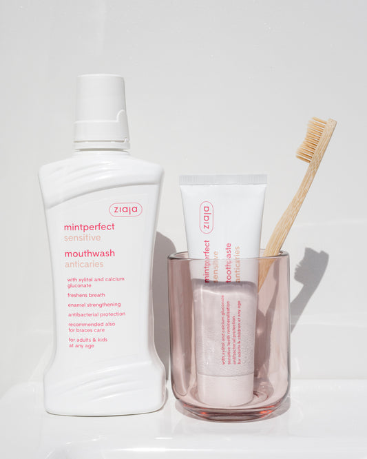 Ziaja Mintperfect Sensitive Anticaries Toothpaste 75ml standing in a pink glass with a bamboo toothbrush, next to a Ziaja Mintperfect Sensitive mouthwash bottle on a bright bathroom surface.