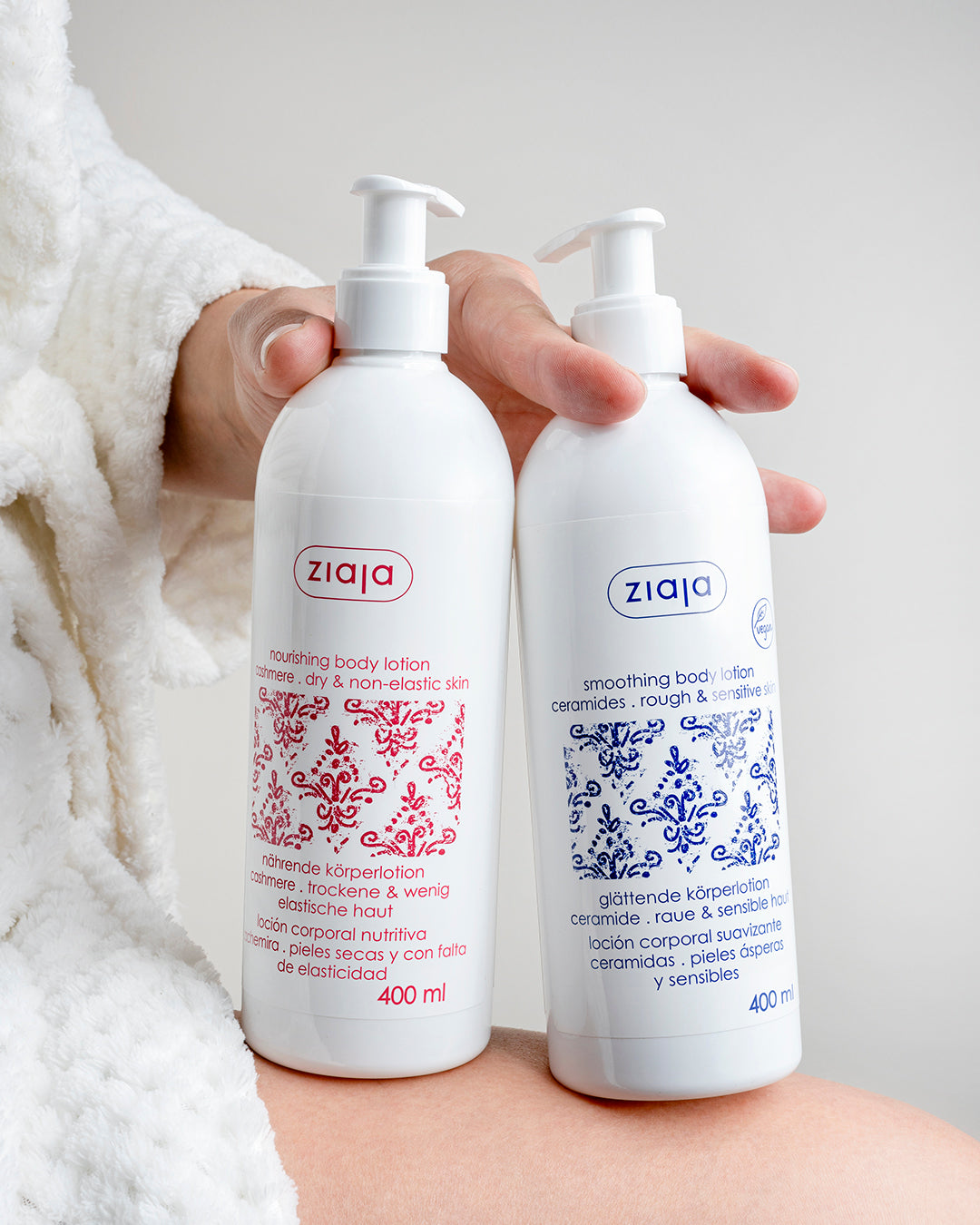 Person in a white bathrobe holding two Ziaja body lotions—Cashmere nourishing body lotion (red label) and Ceramides smoothing body lotion (blue label), 400 ml each.