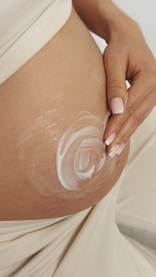 Close-up of a pregnant woman's bare belly with a swirl of white lotion or cream being spread by her hand, wearing a soft cream-colored dress.