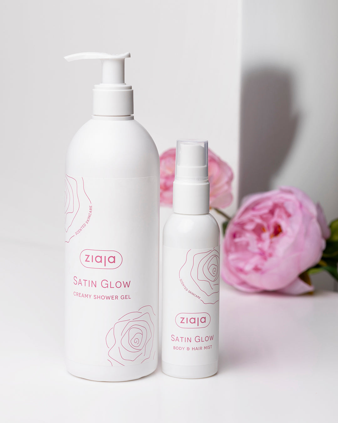 Two Ziaja Satin Glow skincare products with pink flowers in the background on a white surface.