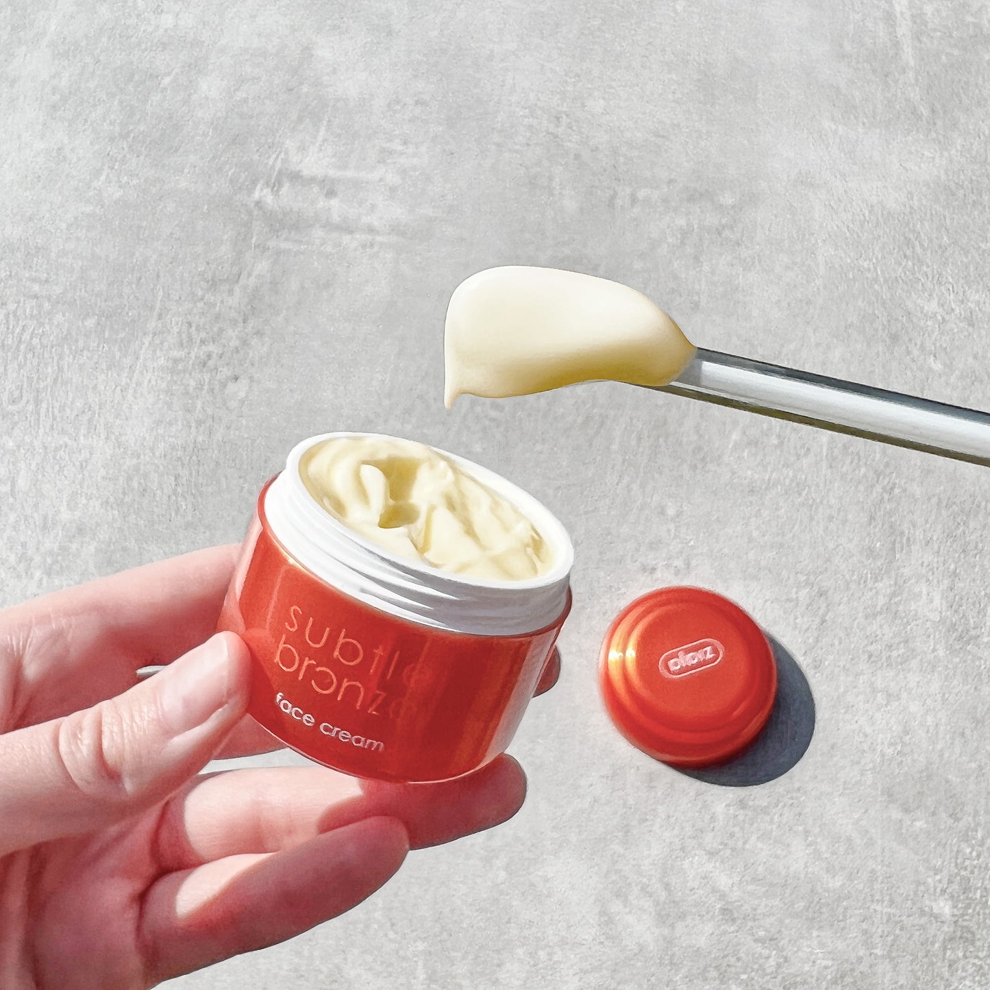Red jar of 'subtil bronz' face cream with a spoonful of cream on a gray background