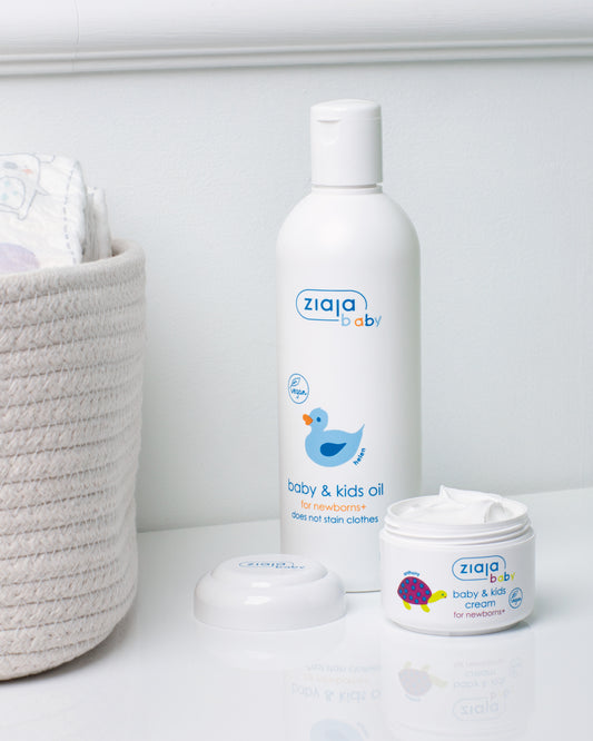 Ziaja Baby & Kids Oil for newborns+ and an open jar of Baby & Kids Cream on a white shelf, with a woven basket in the background.