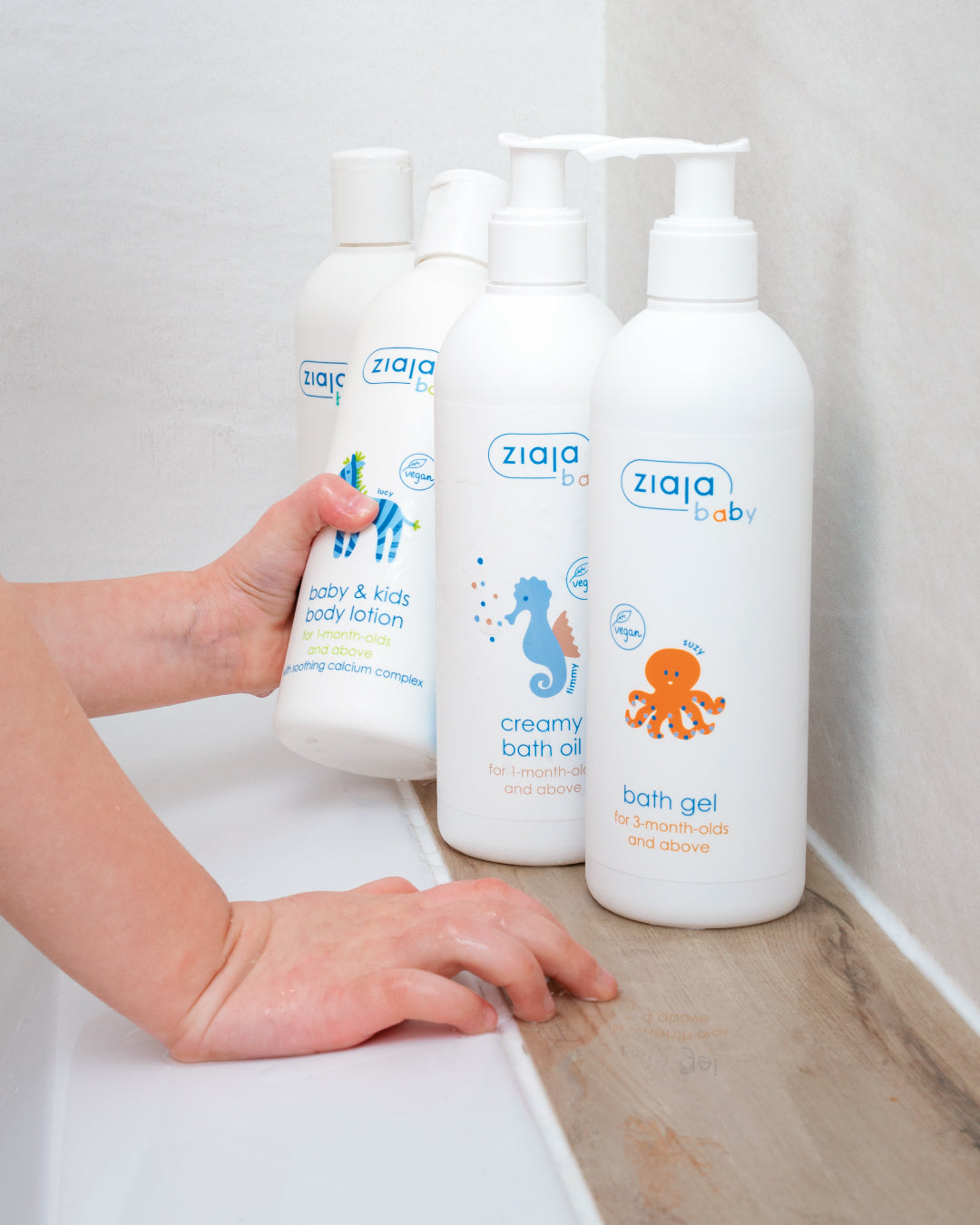 Ziaja Baby Creamy Bath Oil and Ziaja Baby Bath Gel bottles with baby’s hand holding Ziaja Baby & Kids Body Lotion in a bathroom.