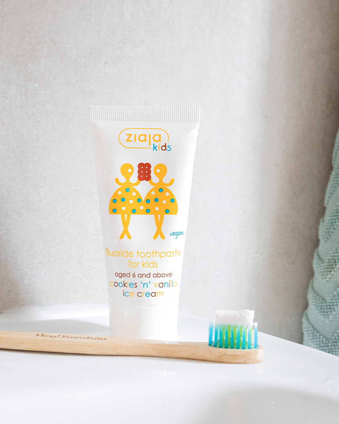 Ziaja Kids fluoride toothpaste (cookies ’n’ vanilla ice cream, ages 6+) standing on a bathroom surface next to a bamboo toothbrush, with a towel in the background.