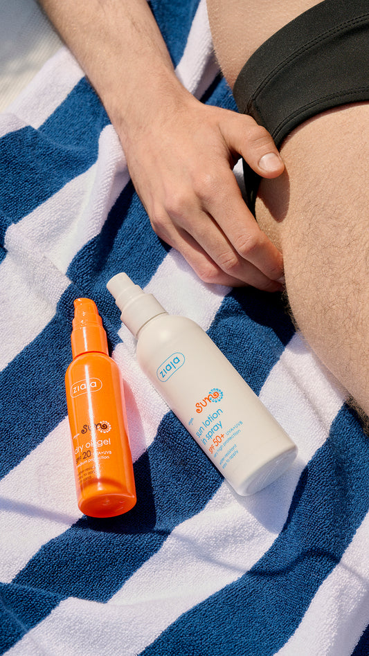 Two Ziaja Sun Moisturising Body Lotion In Spray Spf 50+ 170 Ml and oil unscreen bottles on a striped towel with a person's hand and leg in the background.