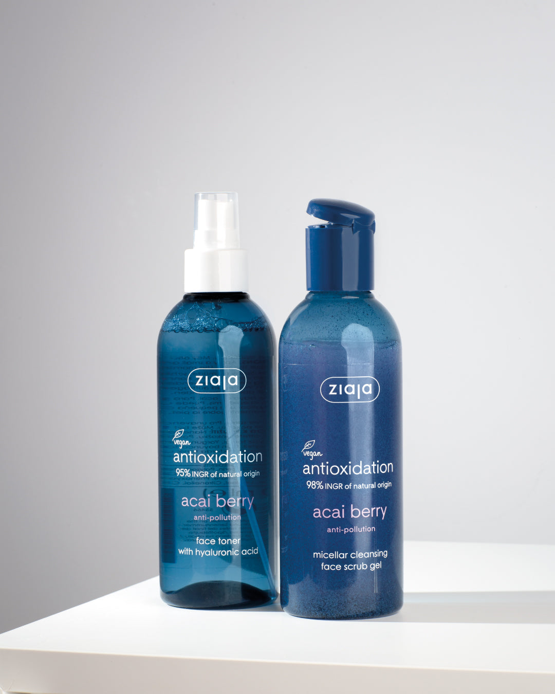 Two blue bottles of Ziaja Acai Berry products agains grey and white ombre background. 