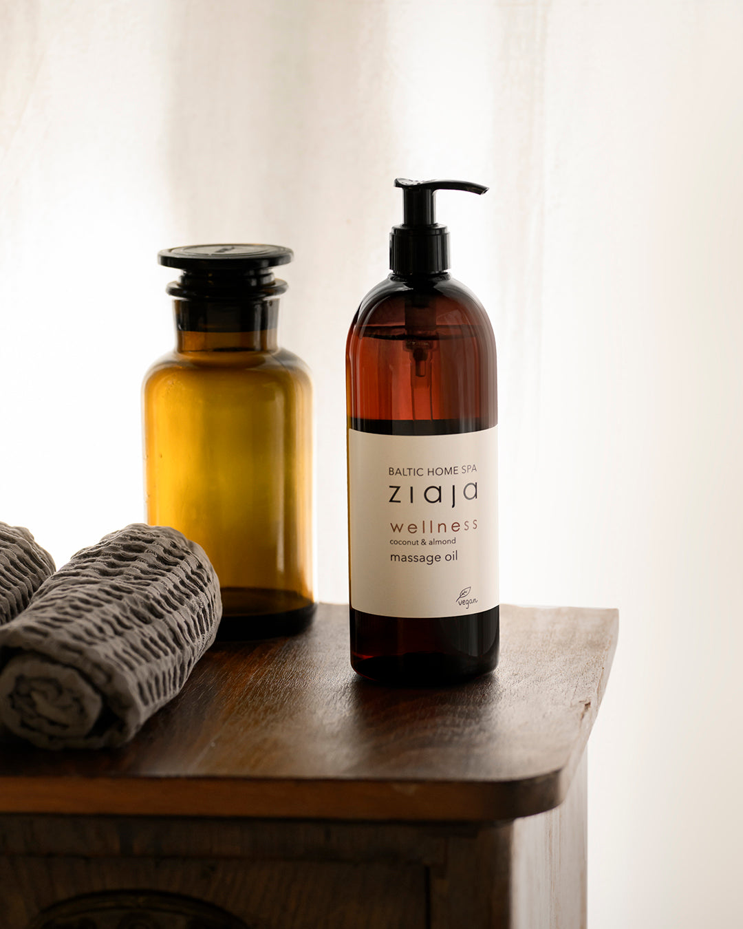 Dark brown pump bottle labeled Ziaja Baltic Home Spa Wellness coconut and almond massage oil placed on a wooden surface next to glass bottles and a folded towel