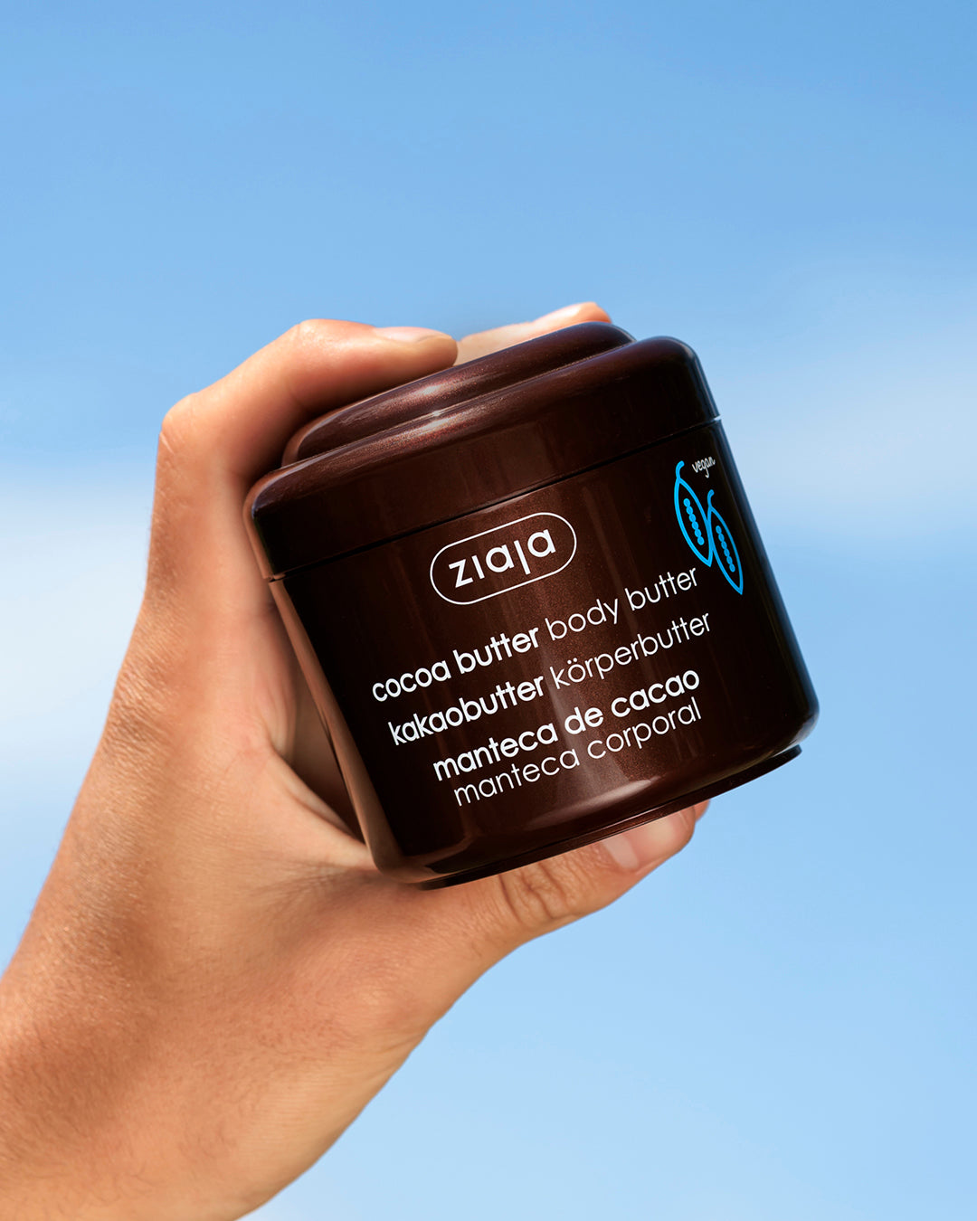 Hand holding a Ziaja cocoa butter body butter container against a blue sky background