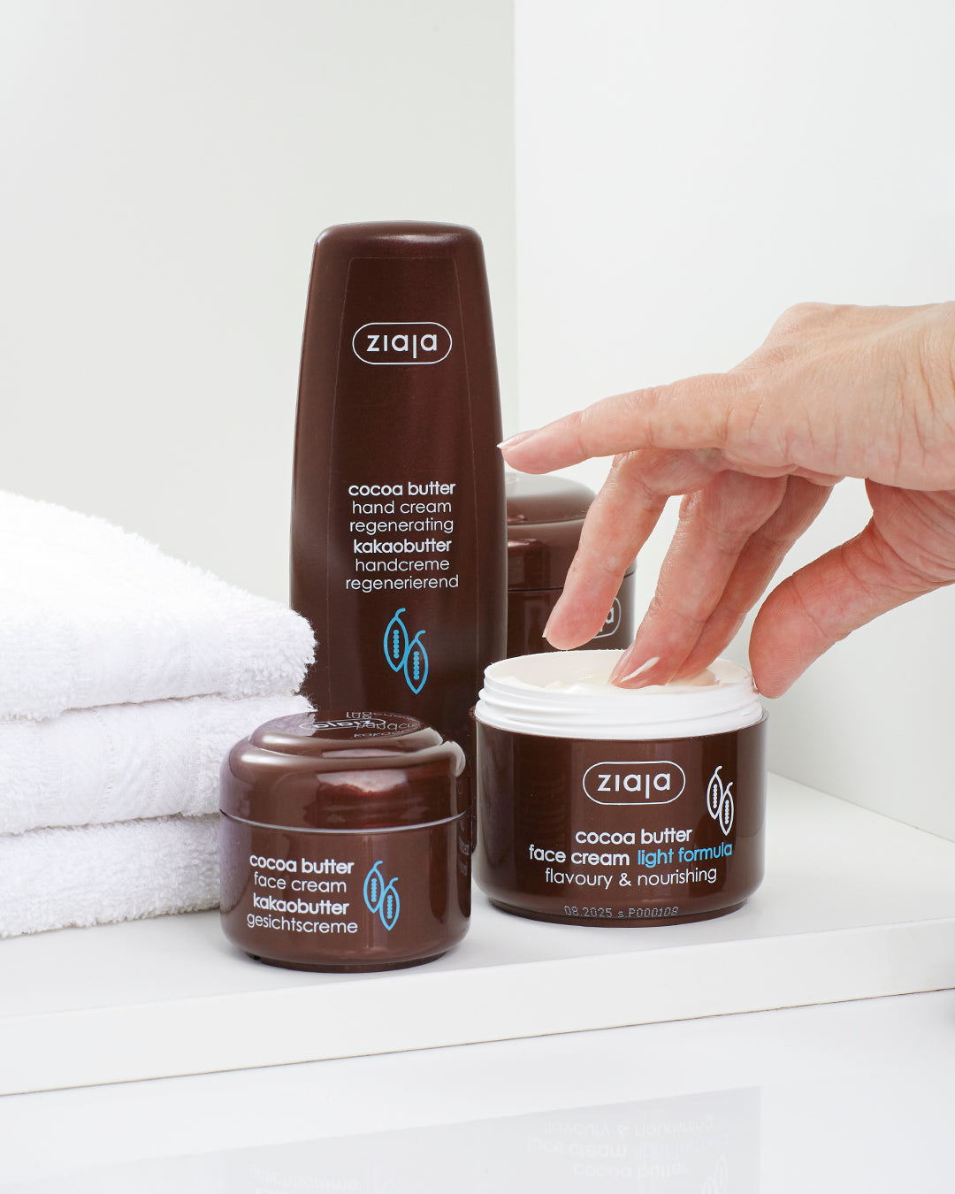 Ziaja cocoa butter hand cream and face cream products in brown packaging placed on a white shelf with a hand reaching into an open jar