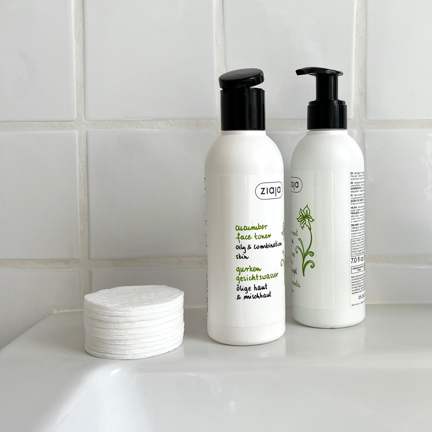 Two white pump bottles of Ziaja Cucumber face care products with black dispensers sit on a white sink counter against a white tiled wall. A stack of white cotton pads is placed beside them.