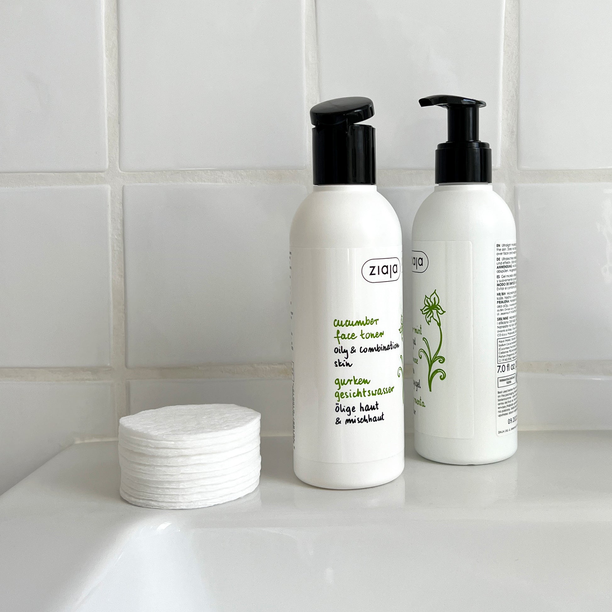 Two white pump bottles of Ziaja Cucumber face care products with black dispensers sit on a white sink counter against a white tiled wall. A stack of white cotton pads is placed beside them.