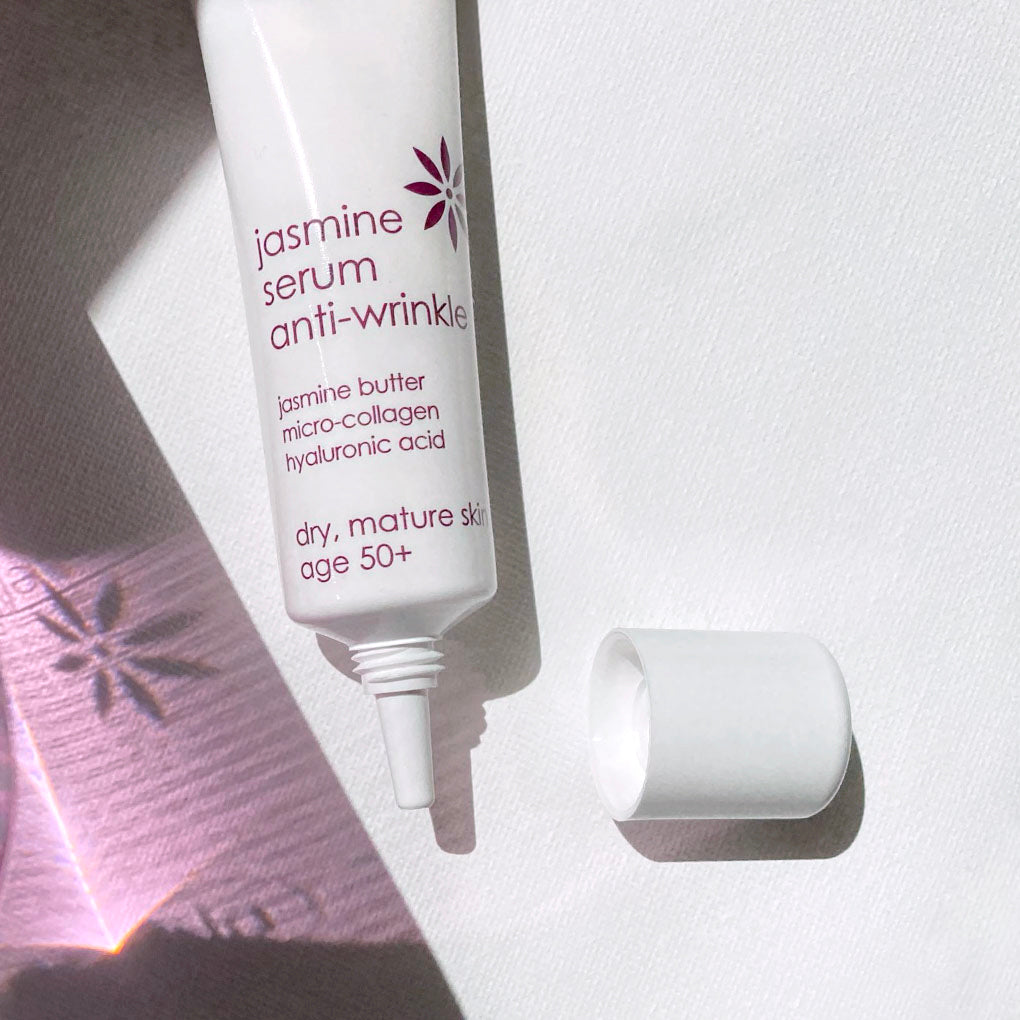 A white squeeze tube of jasmine serum anti-wrinkle cream with its cap removed, placed on a textured white surface with dramatic diagonal shadows and a pink patterned paper accent.