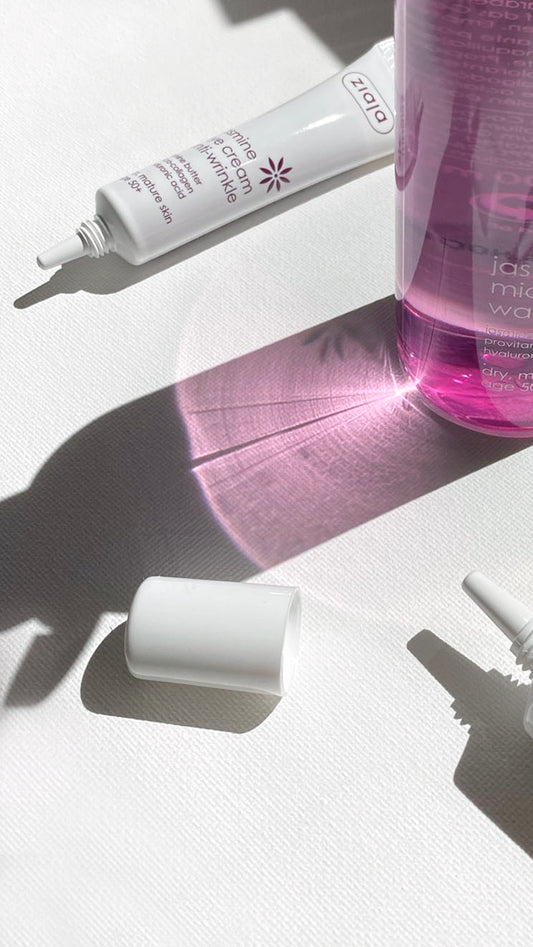 A skincare product arrangement on a white textured surface with strong sunlight and shadows. Visible items include a white squeeze tube labeled 'Ziaja jasmine eye-cream', a translucent pink bottle, a white cap, and a small white applicator tip.