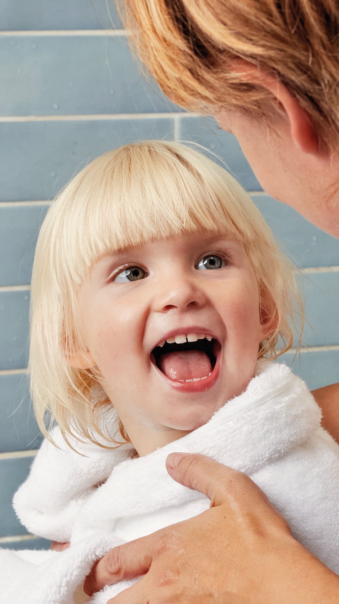 A close-up lifestyle shot of a smiling blonde toddler wrapped in a white towel, looking up at an adult's face against a blue tiled wall.