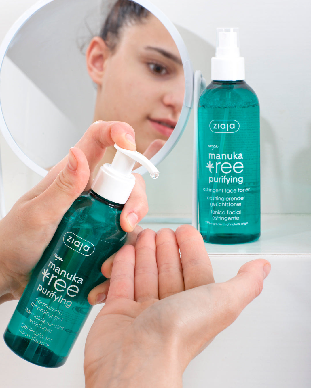 Person holding a bottle of Ziaja manuka tree purifying skincare product in front of a mirror.