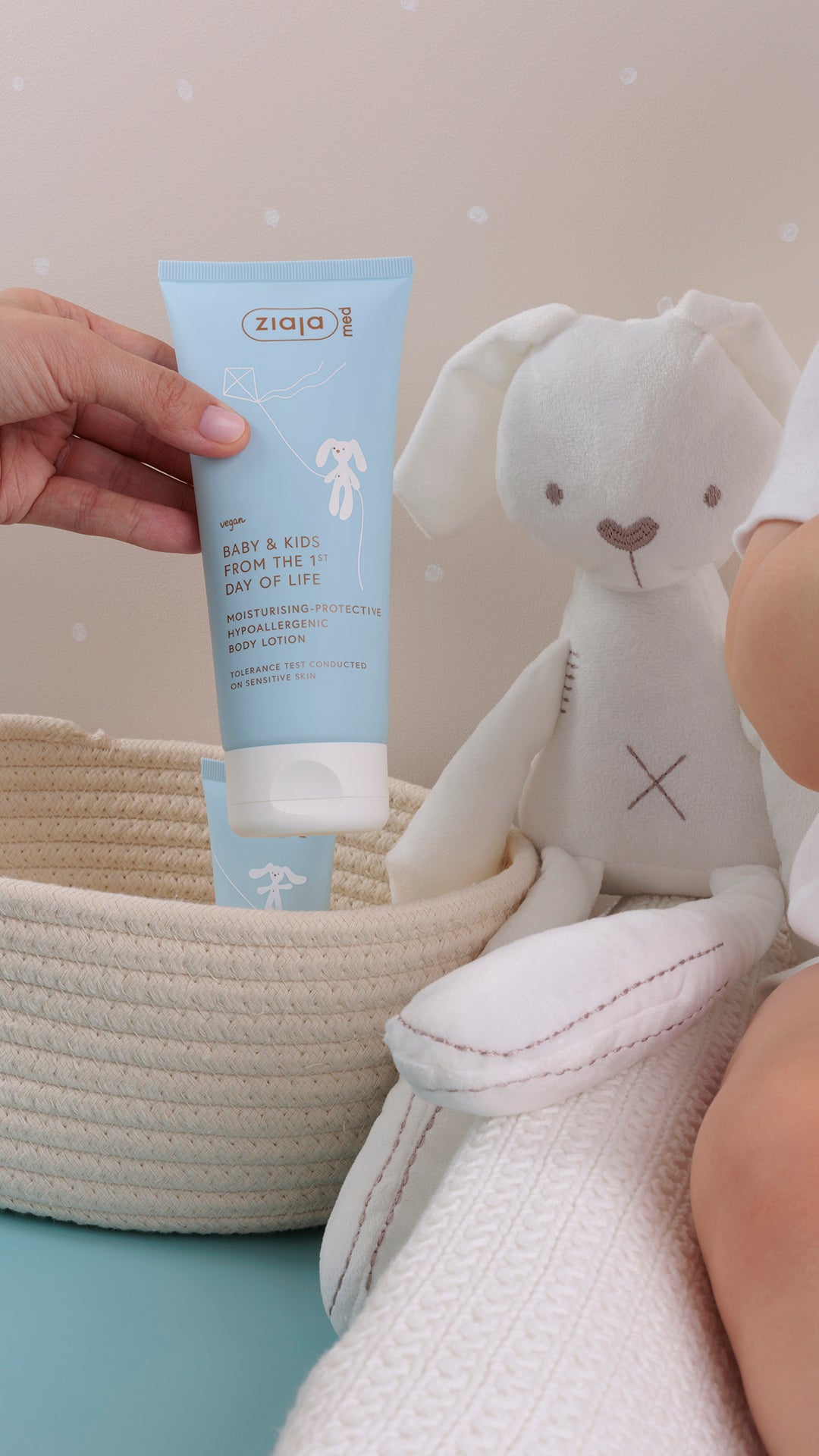 Hand holding Ziaja Med Baby & Kids Hypoallergenic Body Lotion (from the 1st day of life) next to a soft toy and baby on a blanket.