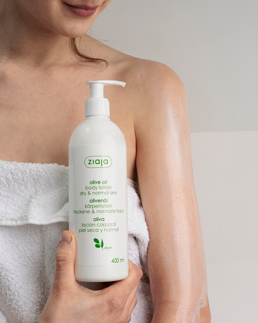 Person holding a bottle of ziaa olive oil body lotion with a white towel draped over their shoulder.