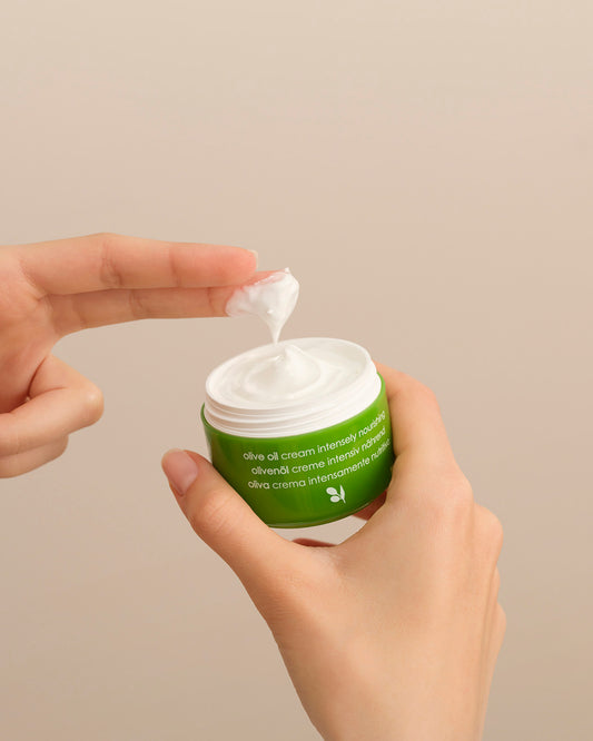 Person applying white Ziaja olive oil cream from a green jar against a plain background
