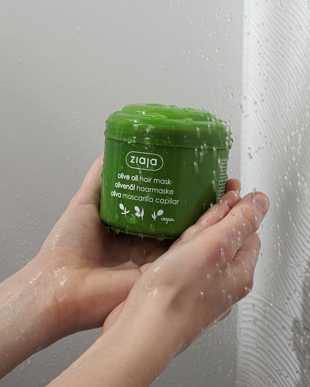 Hand holding a green jar of Ziaja olive oil hair mask against a shower background