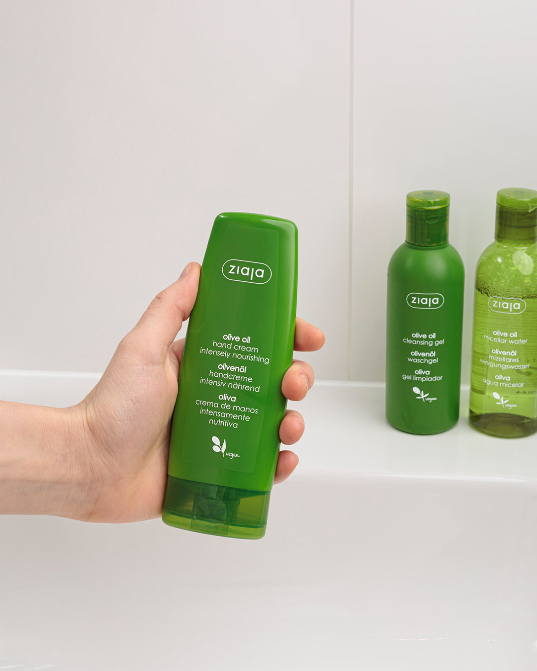 Hand holding a green Ziaja olive oil product with two more bottles in the background on a white surface.