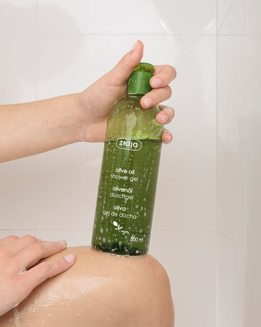 Person applying olive oil shower gel to their skin with a green bottle labeled 'ziaja' on a white tiled wall background.