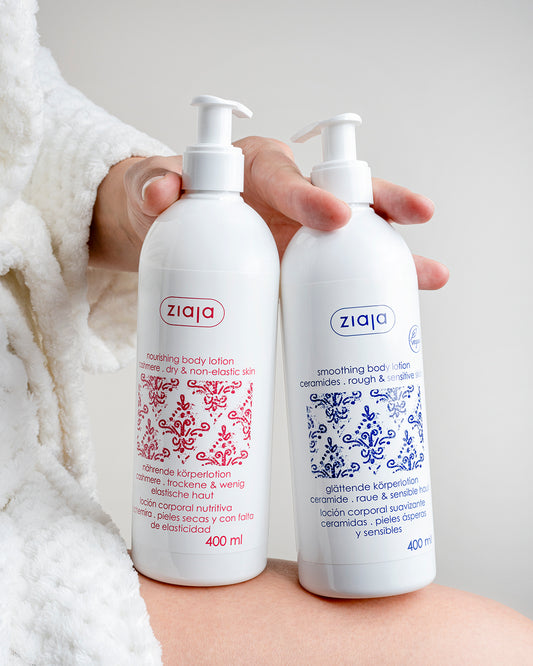 Person in a white bathrobe holding two Ziaja body lotions—Cashmere nourishing body lotion (red label) and Ceramides smoothing body lotion (blue label), 400 ml each.