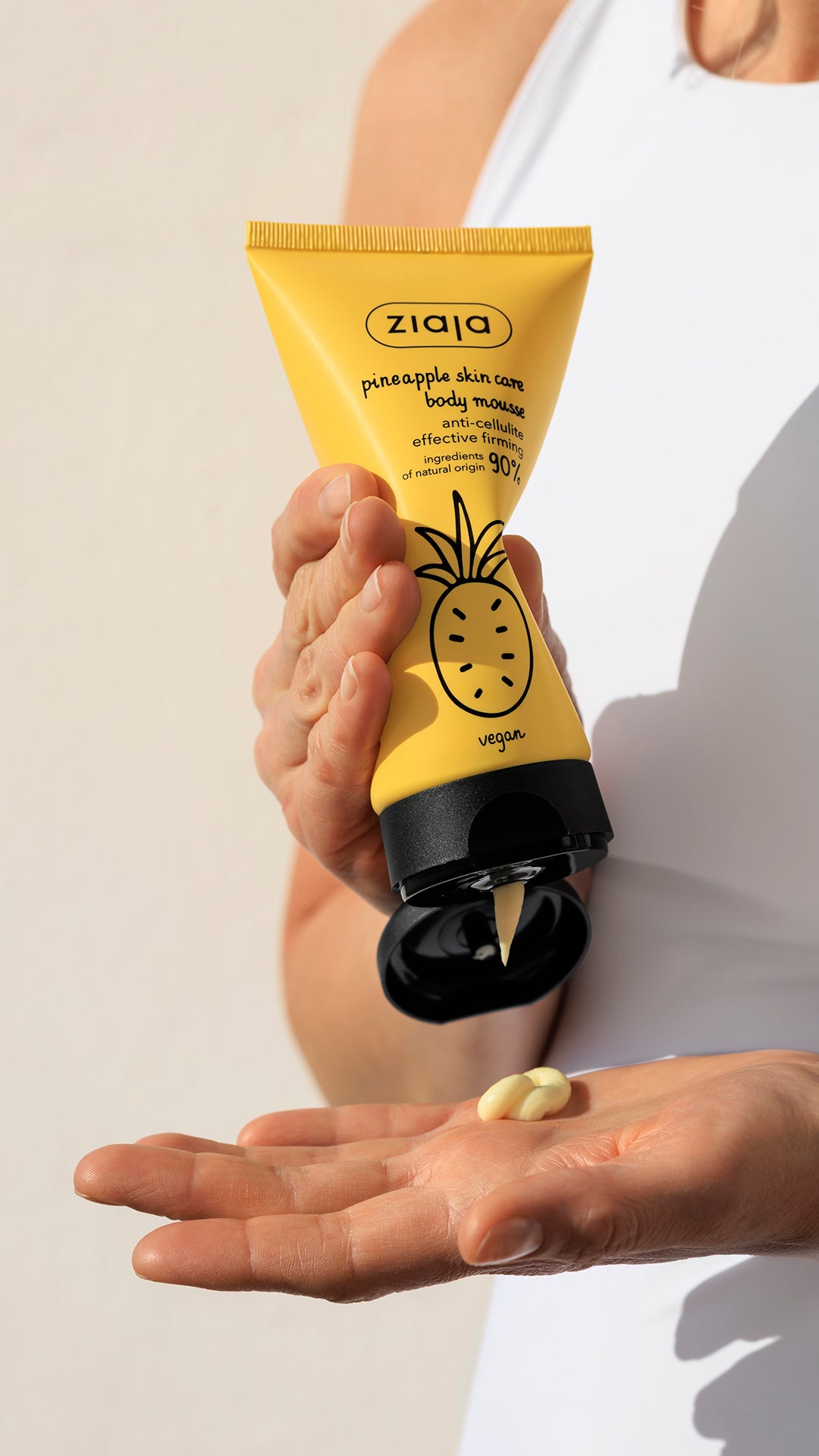 Ziaja Pineapple Anti-Cellulite Body Mousse in a yellow tube, held upside down as a small amount of cream is dispensed onto an open palm in natural sunlight.