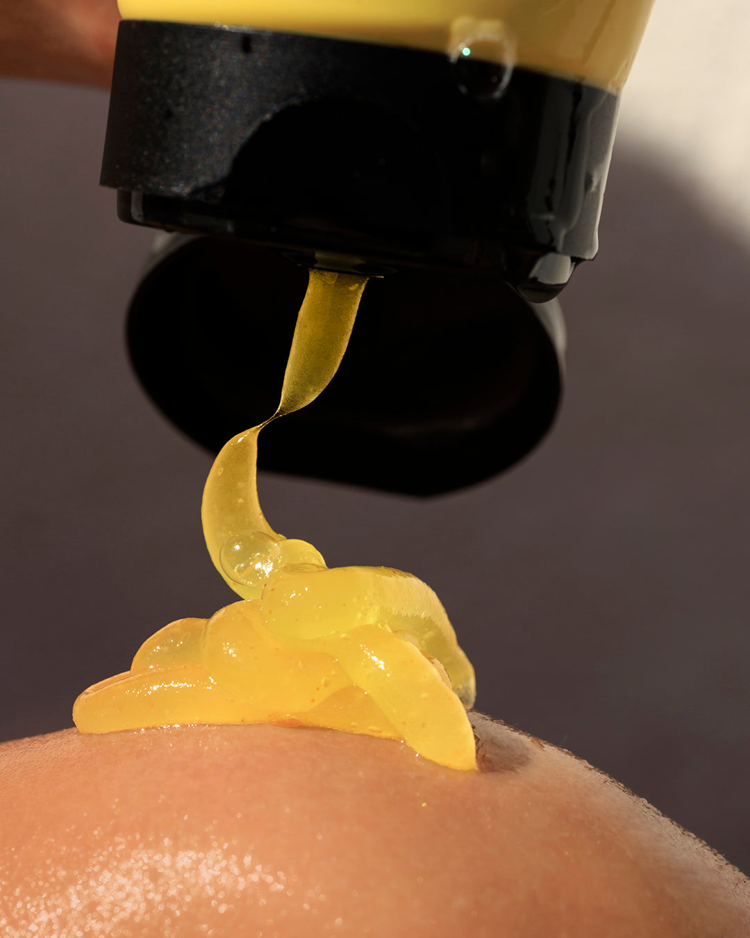 A close-up of a thick, glossy yellow fine grain body scrub being squeezed from a black and yellow tube onto a person's skin.