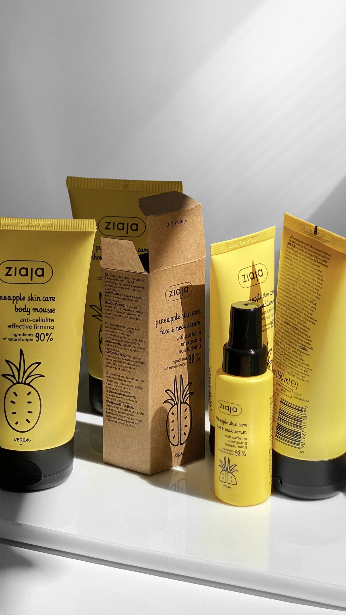 A set of Ziaja pineapple skin care products arranged on a white shelf, including yellow tubes, a brown kraft box, and a small yellow bottle with a black spray cap, against a white background with soft diagonal shadows.