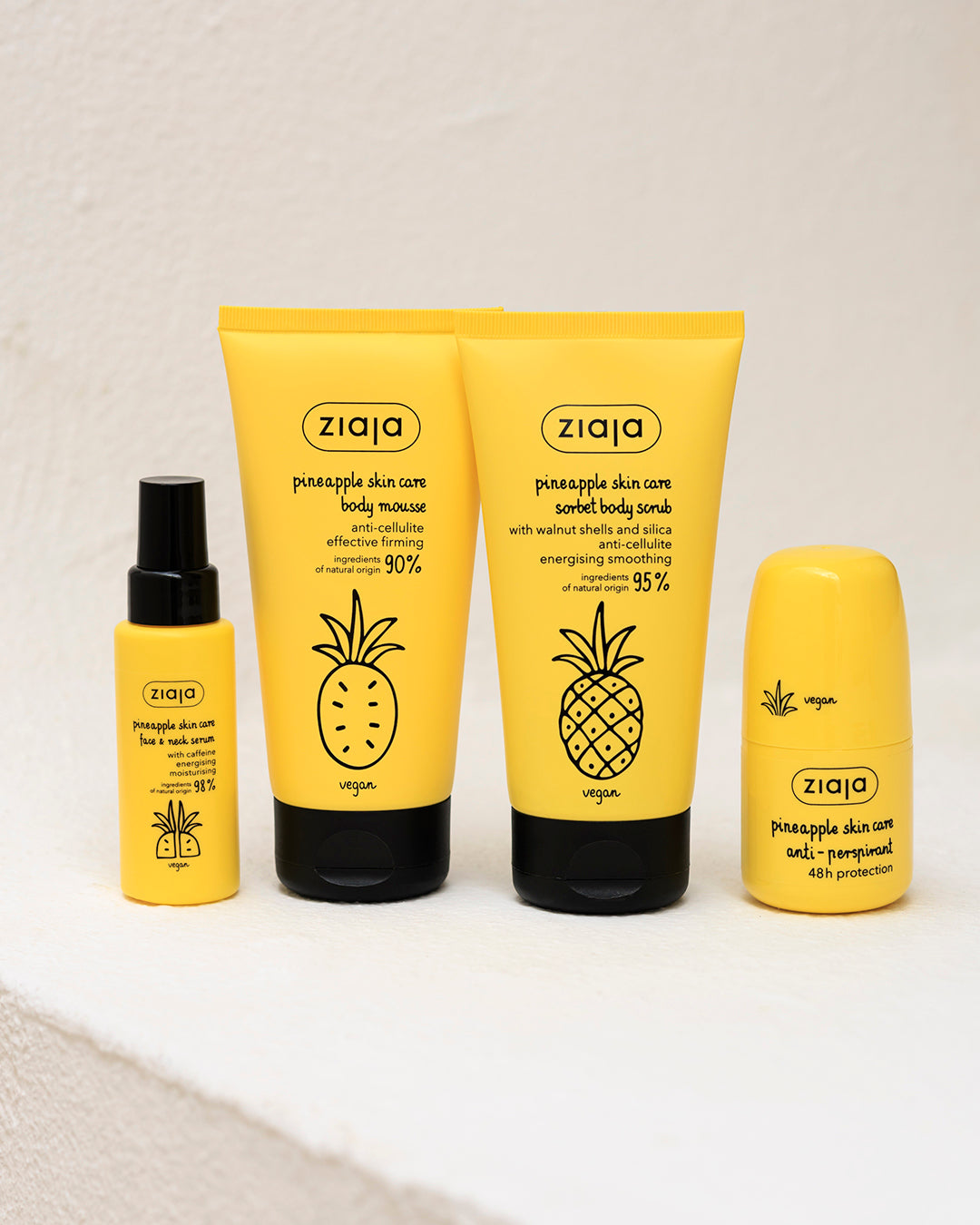 Four Ziaja skincare products in a coordinated yellow and black color scheme, arranged on a white textured surface against a white wall. The set includes a face wash, body wash, body massage, and body scrub.