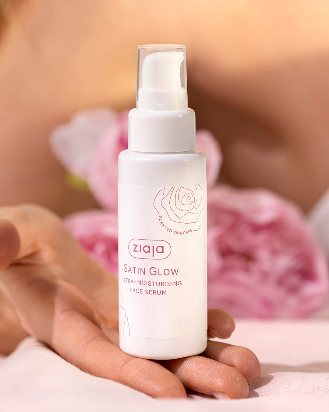Hand holding Ziaja Satin Glow ultra-moisturising face serum bottle with pump, against a soft pink floral background.