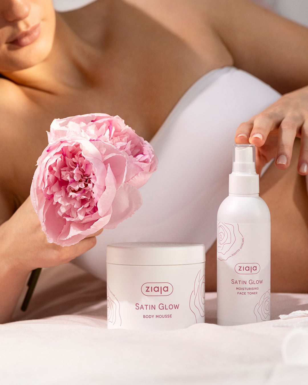 Woman holding a pink flower with Ziaja Satin Glow products on a white background