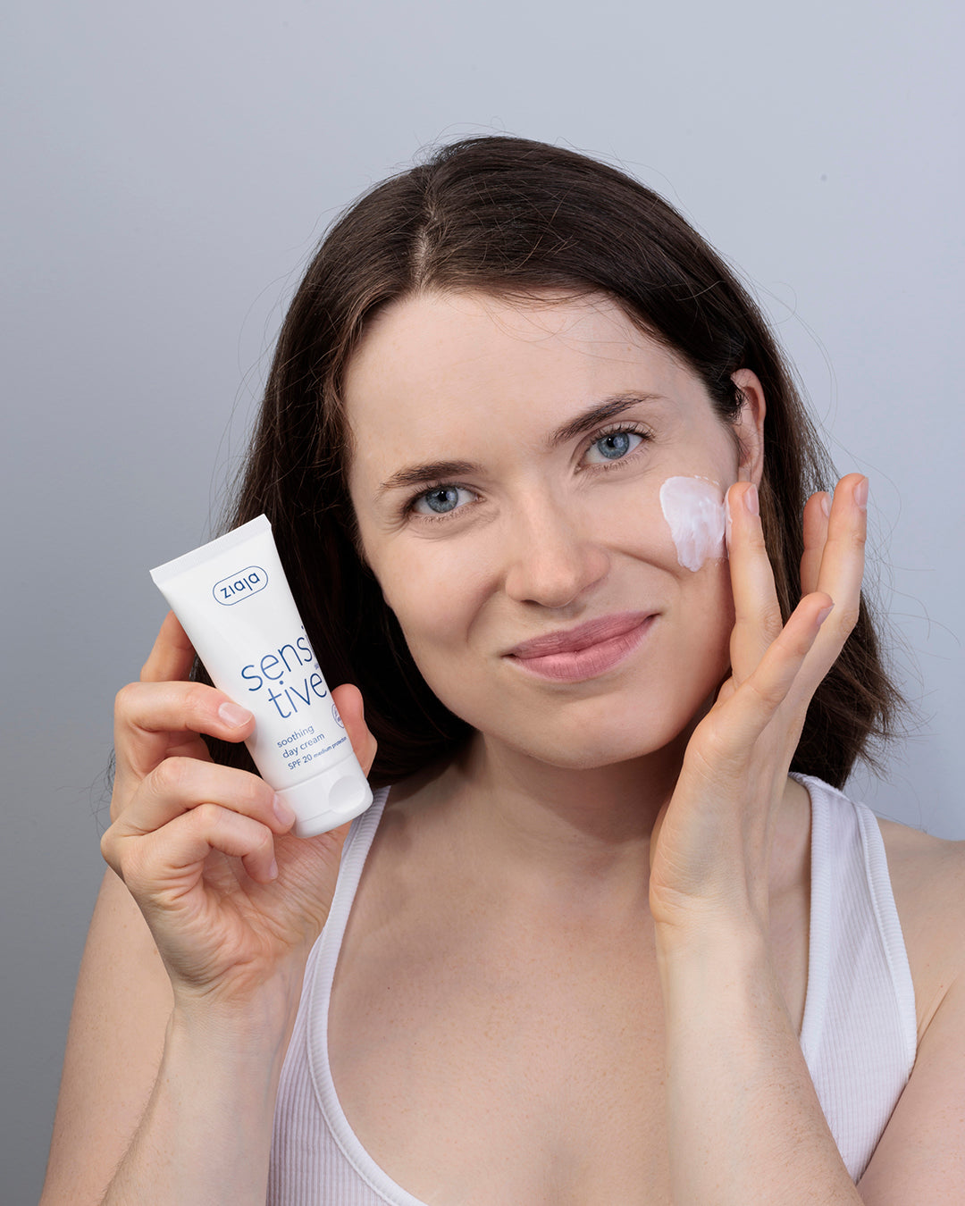 Woman applying Ziaja Sensitive Skin Soothing Day Cream SPF 20 to her cheek while holding the tube.