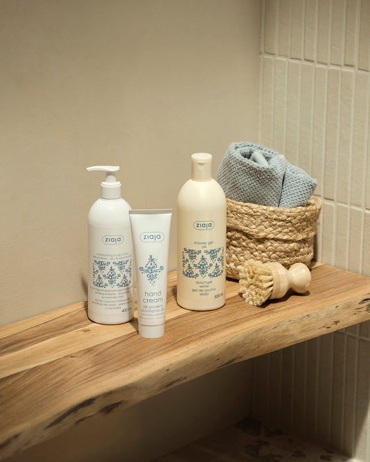 Three white bath and body products from the brand Ziaja Silk Proteins Line are arranged on a rustic wooden shelf next to a woven basket with a folded blue-gray towel and a wooden scrubber, against a beige tiled wall.