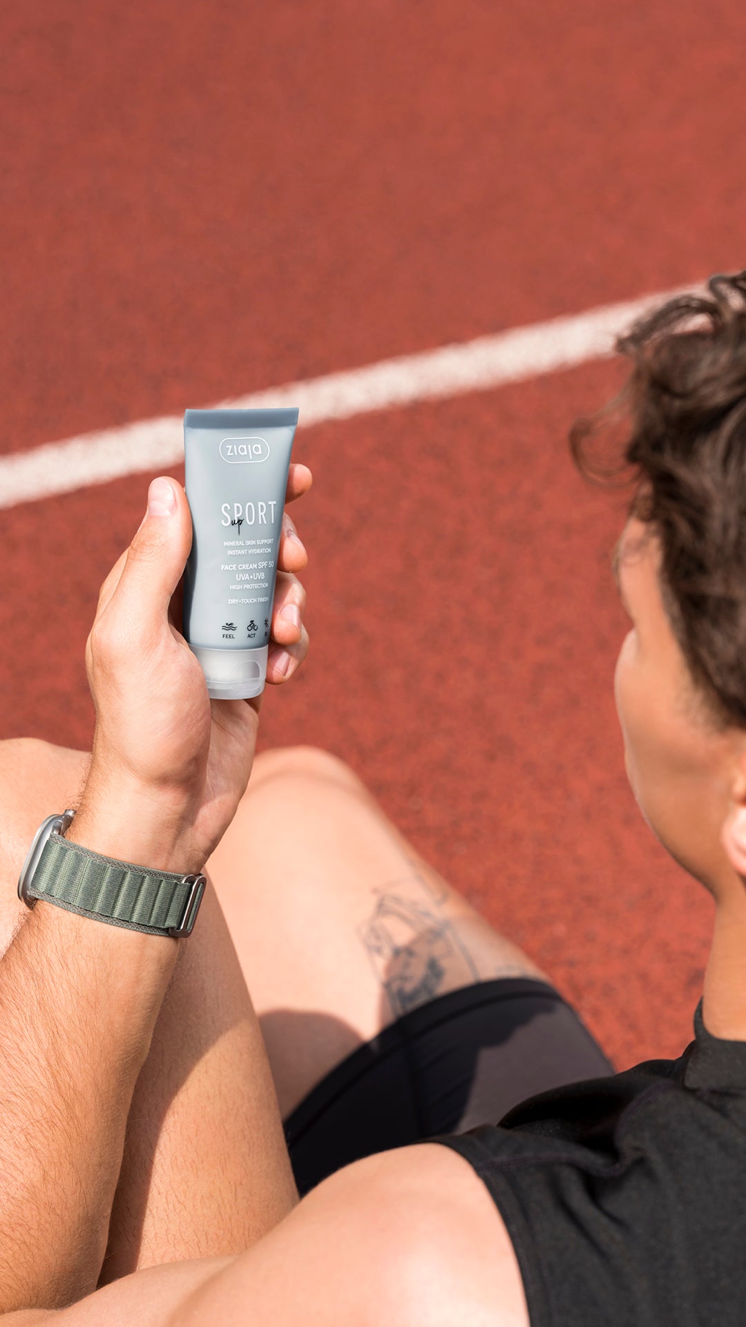 A person sitting on a red running track holds up a grey tube of Ziaja Sport sunscreen. The person is wearing a black athletic shirt and shorts, and the background shows the red track surface with white line markings.