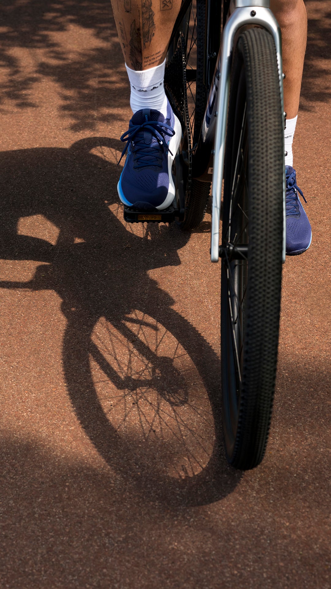 Close-up of a person riding a bicycle on a reddish-brown path, showing blue athletic shoes, white socks, and part of the bike wheel and frame. Strong shadows are cast on the ground.