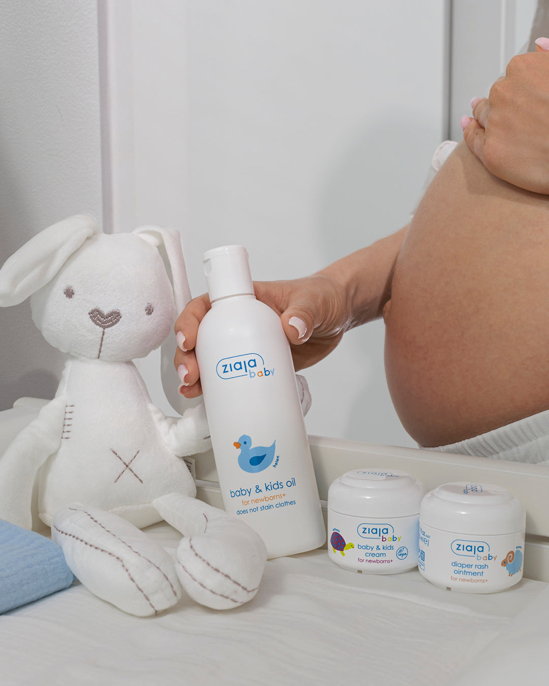 Person holding Ziaja Baby & Kids Oil for newborns+ next to two jars (Baby & Kids Cream and Diaper Rash Ointment) on a baby changing table, with a soft toy bunny.