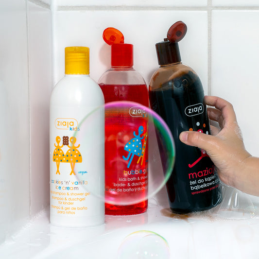 Ziaja Kids bath products in a tiled shower: a white “cookies ’n’ vanilla ice cream” shampoo & shower gel, a red bubble gum kids bath & shower gel, and a black “maZiajki” bubble bath, with a hand holding the black bottle and soap bubbles in the foreground.
