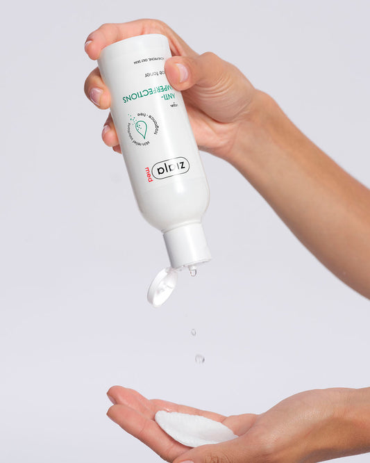 Hand holding a white bottle labeled 'Ziaja Med Anti-imperfections face toner' with product being dispensed onto the palm against a light gray background