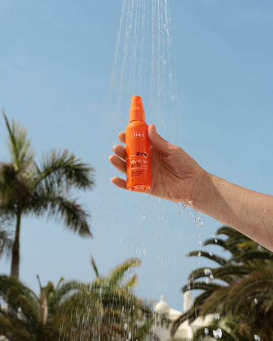 Hand holding Ziaja Sun Dry Oil Gel SPF 20 under running water with palm trees in the background