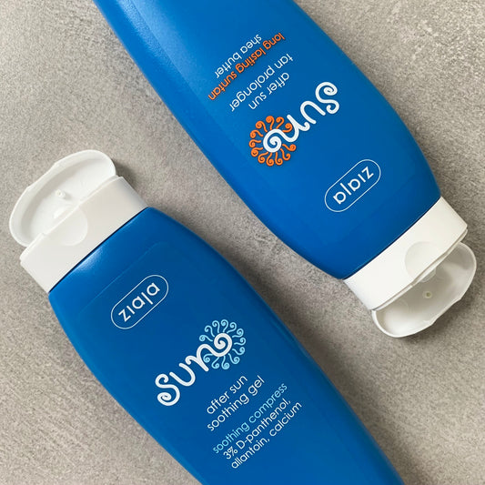 Two Ziaja Sun blue bottles (after sun soothing gel and tan prolonger) on sand, caps open.