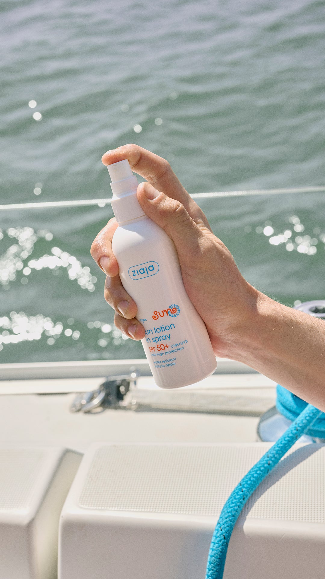 Hand holding a Ziaja Sun Moisturising Body Lotion In Spray Spf 50+ 170 Ml bottle of sunscreen spray with a blurred water background